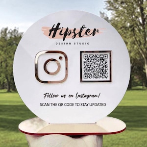 Peut inclure: Une pancarte ronde blanche avec le texte "Hipster Design Studio" en lettres noires. La pancarte présente également un logo Instagram rose et un code QR noir et blanc avec le texte "Suivez-nous sur Instagram ! Scannez le code QR pour rester à jour".