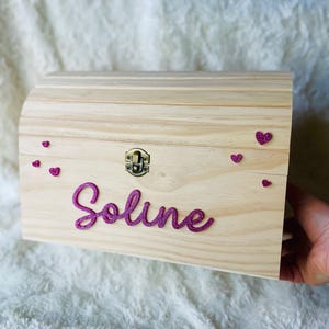 Könnte beinhalten: Eine Holzkiste mit gewölbtem Deckel, verschlossen mit einem Metallverschluss. Der Name "Soline" ist in glitzerndem Pink geschrieben. Kleine rosa Herzdekorationen zieren die Kiste. Ideal zum Aufbewahren von Andenken.