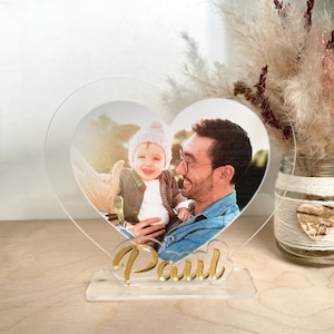 Pode incluir: Um porta-retratos de coração de acrílico transparente com uma foto de um pai e uma criança. O porta-retratos tem um suporte dourado e o nome "Paul" está escrito em letras douradas no suporte.