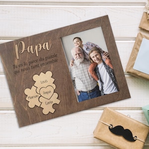 May include: A wooden photo frame with a brown background and a photo of a family. The frame has a cutout puzzle piece design with the words "Papa", "Maman", "Louis", "Carla" and "Papa" inscribed on the pieces. The text "Tu es la pièce du puzzle qui nous tient ensemble" is also inscribed on the frame.