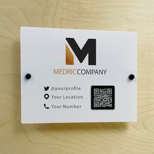 Könnte beinhalten: Ein weißes Schild mit einem schwarzen M-Logo und dem Text "MEDRICCOMPANY". Unter dem Logo befinden sich Social-Media-Symbole und ein QR-Code. Der Text lautet "@yourprofile", "Your Location" und "Your Number".