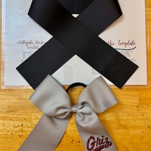 Puede incluir: Un lazo de animadora gris con un logotipo "Griz" de purpurina marrón. El lazo está atado con una cinta negra y una goma elástica negra para el pelo.