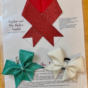 May include: A template for making a keychain or mini hair bow using 14 inches of 1.5 inch ribbon. The template shows a red glitter ribbon folded into a bow shape with instructions for placement of a decal. There are two finished bows, one green glitter and one white glitter, with a keychain ring and a hair tie.