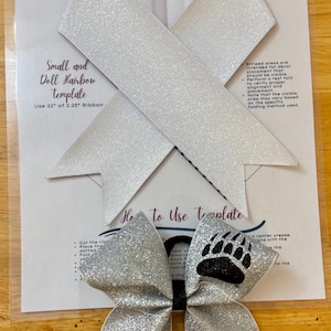 May include: A silver glitter hair bow with a black bear paw print and the word "GRIZZLIES" in black text. The bow is on a white background with a template for making a hair bow. The template includes instructions for cutting and folding a 22 inch ribbon.