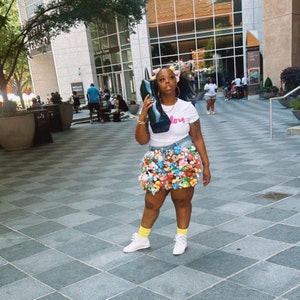 Puede incluir: Una persona lleva una camiseta blanca con la palabra "Love" impresa, una falda vaquera con adornos coloridos y zapatillas blancas. La persona lleva un bolso de mano negro.