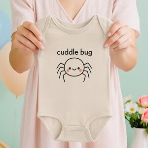 May include: A beige baby onesie with the words "cuddle bug" printed in black. Below the text is a cartoon spider with a smiling face. The onesie is held up by a person.