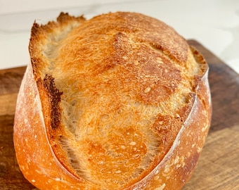 Puede incluir: Una hogaza de pan de masa madre recién horneada con una corteza dorada y una forma ligeramente irregular.