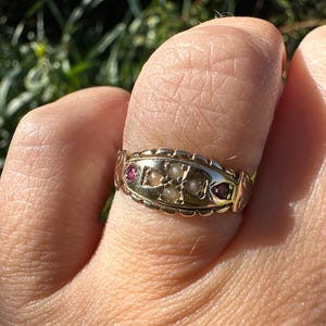 Peut inclure: Une bague en or avec une bande décorative ornée de petites perles et de pierres précieuses rouges. La bague est portée au doigt, la lumière du soleil mettant en valeur les détails du bijou.