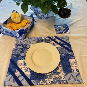 Puede incluir: Un mantel individual azul y blanco con un plato blanco encima. El mantel individual está hecho de tela y tiene un diseño de patchwork. Hay una copa de vino tinto y un posavasos azul y blanco en la mesa.