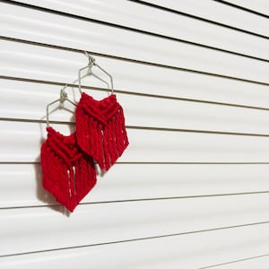 May include: A pair of red macrame earrings with geometric shapes and fringe. The earrings are hanging against a white background.