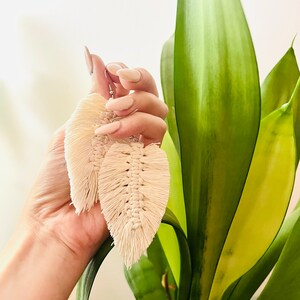 May include: A pair of cream-colored feather-shaped earrings made of macrame. The earrings are hanging from a person's hand.