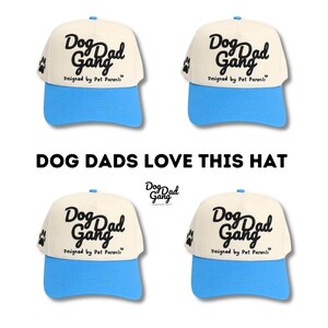 May include: Cream-colored baseball cap with a light blue brim and top button. The cap features the text "Dog Dad Gang" in black script, with a paw print on the side. The phrase "Designed by Pet Parents" is also printed on the cap.