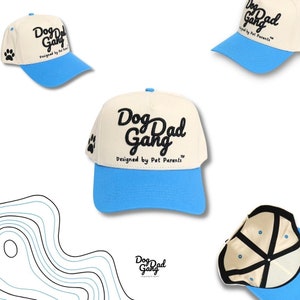 May include: Cream-colored baseball cap with a light blue brim. The cap features the text "Dog Dad Gang" in black script, with "Designed by Pet Parents" below. A paw print is on the side.
