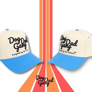 May include: Two baseball caps with a cream-colored crown and a light blue brim. The caps have the text "Dog Dad Gang" in black script. The phrase "Designed by Pet Parents" is below the main text. The background has orange and red stripes.