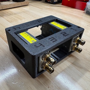 May include: A black, textured, rectangular 3D-printed frame housing two electronic devices labeled Fido Set 05. The frame features four gold-colored BNC connectors on one side and embossed text on the top surface, providing a secure mount.
