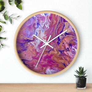 May include: A round wall clock with a wooden frame and a colourful abstract design in shades of pink, purple, and gold. The clock face has white hands and a white background.