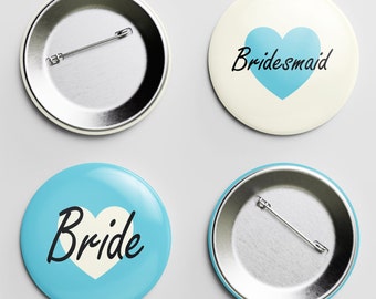 May include: Four round pinback buttons. Two buttons have a silver metal back with a safety pin. One button is light blue with a white heart and the word "Bride" in black. Another button is off-white with a blue heart and the word "Bridesmaid".