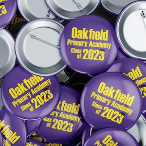 Könnte beinhalten: Ein Stapel runder, violetter Knöpfe mit dem Text "Oakfield Primary Academy Class of 2023" in gelben Buchstaben.
