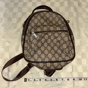 May include: A small, brown backpack with a repeating pattern. The backpack has a top handle with a green and red stripe, brown straps, and a zippered front pocket. The bag is approximately 10 inches wide.