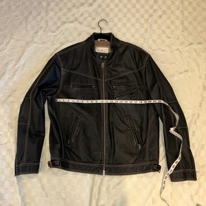 Könnte beinhalten: Schwarze Lederjacke mit Reißverschluss, mehreren Taschen und braunen Nähten. Die Jacke hängt an einem schwarzen Kleiderbügel und hat einen braunen Kragen. Ein Maßband liegt über der Jacke.