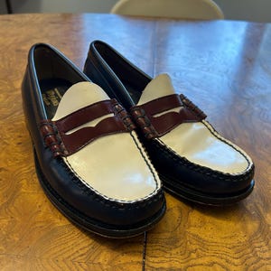 May include: Pair of two-tone leather penny loafers. The shoes feature a navy blue body, white vamp, and a burgundy strap. The soles are black with white stitching. The shoes are displayed on a wooden surface.