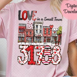May include: A light pink t-shirt featuring a graphic design with the words "LOVE in a Small Town" above a street scene and the numbers "31738". The word "LOVE" is in a mix of fonts and patterns, including black glitter and gingham.