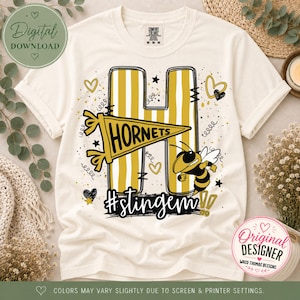 Peut inclure: Un t-shirt couleur crème arborant une grande lettre H à rayures verticales jaunes et blanches. Un fanion jaune avec le texte Hornets est superposé sur la lettre. Un frelon de dessin animé, des petits cœurs, des étoiles et le hashtag #stingem.