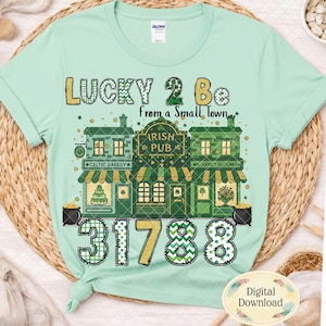 May include: A pale green t-shirt with a graphic design. The design includes the text "LUCKY 2 Be from a Small Town", an Irish pub, a bakery, and the numbers "31788". The design is in a festive style. Digital download.