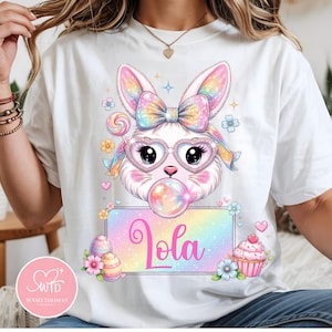 May include: White t-shirt featuring a colorful cartoon bunny with heart-shaped glasses blowing a bubble. The bunny has a rainbow bow and is surrounded by Easter-themed graphics, including an egg, cupcake, and the name "Lola".