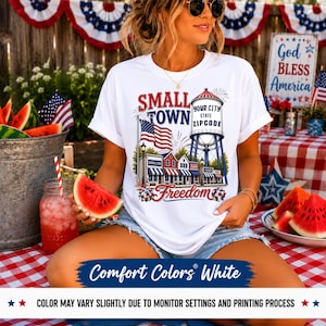 May include: A white t-shirt featuring a graphic print of a small town scene with a water tower, an American flag, and storefronts. The text reads Small Town, Freedom, and Your City State Zipcode. The design includes red, white, and blue colours.