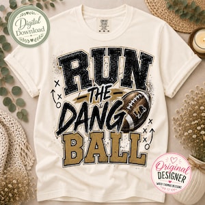 May include: A cream-colored short-sleeve t-shirt featuring a graphic print with the text RUN THE DANG BALL in black and gold distressed lettering. The design includes a brown football illustration and small white play diagrams on the front.
