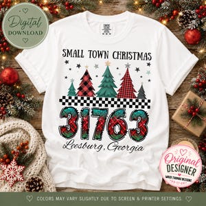 Op de afbeelding: Een wit T-shirt met korte mouwen met de tekst Small Town Christmas, een rij van vijf gestileerde kerstbomen, een zwart-wit geblokte lijn, het getal 31763 in een rood en groen ruitpatroon en de tekst Leesburg, Georgia.