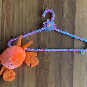 May include: A pink, blue, and purple fabric hanger with a flower embellishment. A plush orange crab toy is hanging from the hanger.