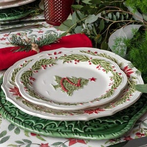 Op de afbeelding: Een feestelijke kersttafelsetting met witte borden met een kerstboomontwerp, rode servetten en groene placemats. Heldere wijnglazen en rood gedessineerde glazen zijn ook aanwezig, omringd door groen en poinsettia's.
