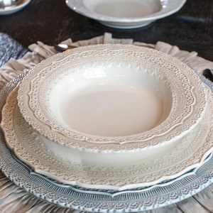Puede incluir: Un juego de plato hondo y plato llano de cerámica color crema con un intrincado patrón en relieve similar al encaje a lo largo de los bordes festoneados. El diseño incluye un centro liso y brillante con un borde decorativo texturizado.