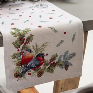 Könnte beinhalten: Ein weißer Tischläufer mit einem winterlichen Design mit zwei rotbrüstigen Vögeln, die auf einem Zweig mit roten Beeren sitzen. Der Hintergrund ist mit Tannennadeln und roten Beeren übersät.