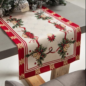 May include: A Christmas table runner with a cream-coloured centre and a red border. The runner features a repeating pattern of bells, poinsettias, and ribbons. The border has a snowflake design. The runner is placed on a grey table.