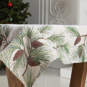 Puede incluir: Un mantel blanco con un patrón de agujas de pino verdes y piñas. El mantel está drapeado sobre una mesa de madera.