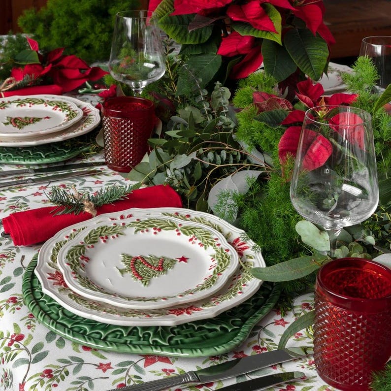 Op de afbeelding: Een feestelijke kersttafelsetting met witte borden met een kerstboomontwerp, rode servetten en groene placemats. Heldere wijnglazen en rood gedessineerde glazen zijn ook aanwezig, omringd door groen en poinsettia's.