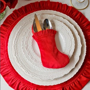 Op de afbeelding: Een rode fluwelen bestekhouder in de vorm van een kerstsok met een geplooide bovenrand, met daarin een zilveren mes en lepel. Het ligt op een wit bord met een gedetailleerde kanten rand, op een grote rode geplooide onderbord van stof.