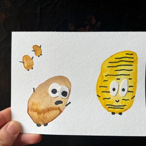 May include: Watercolor illustration of three potato-shaped characters with googly eyes on a white background. One is brown, another is yellow with black stripes, and the other three are small and brown. The artwork is on a white card.