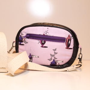 May include: A light pink crossbody bag with a black leather trim and a cream-colored strap. The bag features a purple zipper and is decorated with illustrations of potion bottles, butterflies, and figures. The bag is designed for carrying essentials.