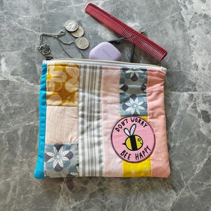 May include: A patchwork zippered pouch with a "Don't Worry Bee Happy" patch. The pouch is filled with a comb, coins, a necklace, and a small purple item. The pouch has a variety of fabric patterns in shades of blue, pink, yellow, and gray.
