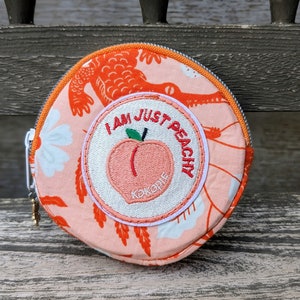 以下が含まれることがあります： ピンクとオレンジの丸い布製の小銭入れで、白い刺繍のワッペンには「I am just peachy」と、その中に桃と「Kokoolie」の文字が書かれています。財布にはジッパーと小さな金のチャームが付いています。