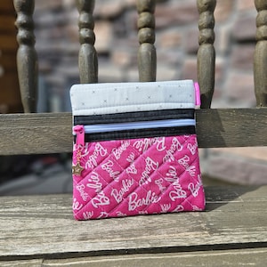Puede incluir: Una bolsa de cremallera acolchada de color rosa con fondo blanco y la palabra "Barbie" repetida en letras blancas. La bolsa tiene una cremallera negra y un charm de estrella dorada.