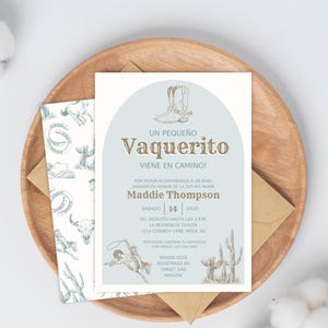 May include: A baby shower invitation with a cowboy theme. The invitation features the text "Un pequeño Vaquerito viene en camino!" in brown and a light blue arch with a cowboy boot illustration. The invitation is on a wooden plate with a cotton branch.