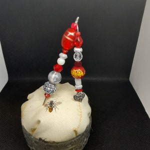 Puede incluir: Un alfiletero de cuentas rojas y plateadas con un dije de corazón y un diseño de abeja en la tela.