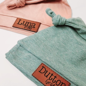 May include: Two knotted hats in soft pastel colors, one light pink and one sage green. Each hat features a folded brim and a rectangular brown leather-like patch stitched to the front. The pink hat displays the text Luna May, while the green hat displays the text Dutton.