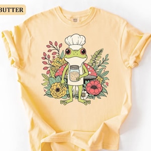 May include: Butter-colored t-shirt featuring a cartoon frog chef holding a jar of spices, surrounded by flowers and mushrooms. The frog wears a chef's hat and apron. The word "BUTTER" is in a yellow rectangle in the upper left corner.