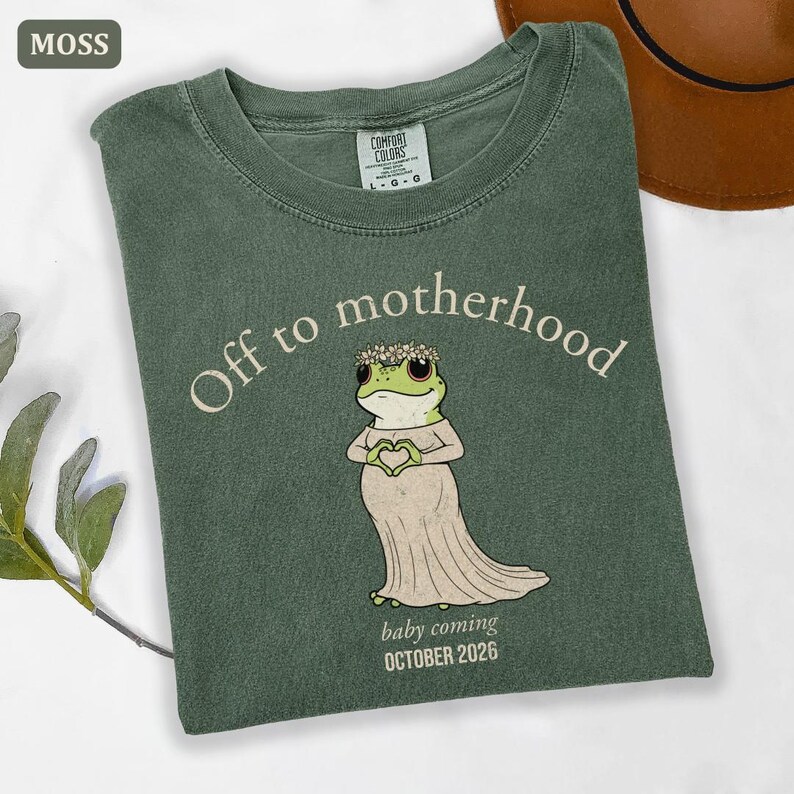 May include: Moss green t-shirt with the text "Off to motherhood" and a cartoon frog wearing a floral crown and dress, holding a heart shape over its belly. The text "baby coming OCTOBER 2026" is below the frog.